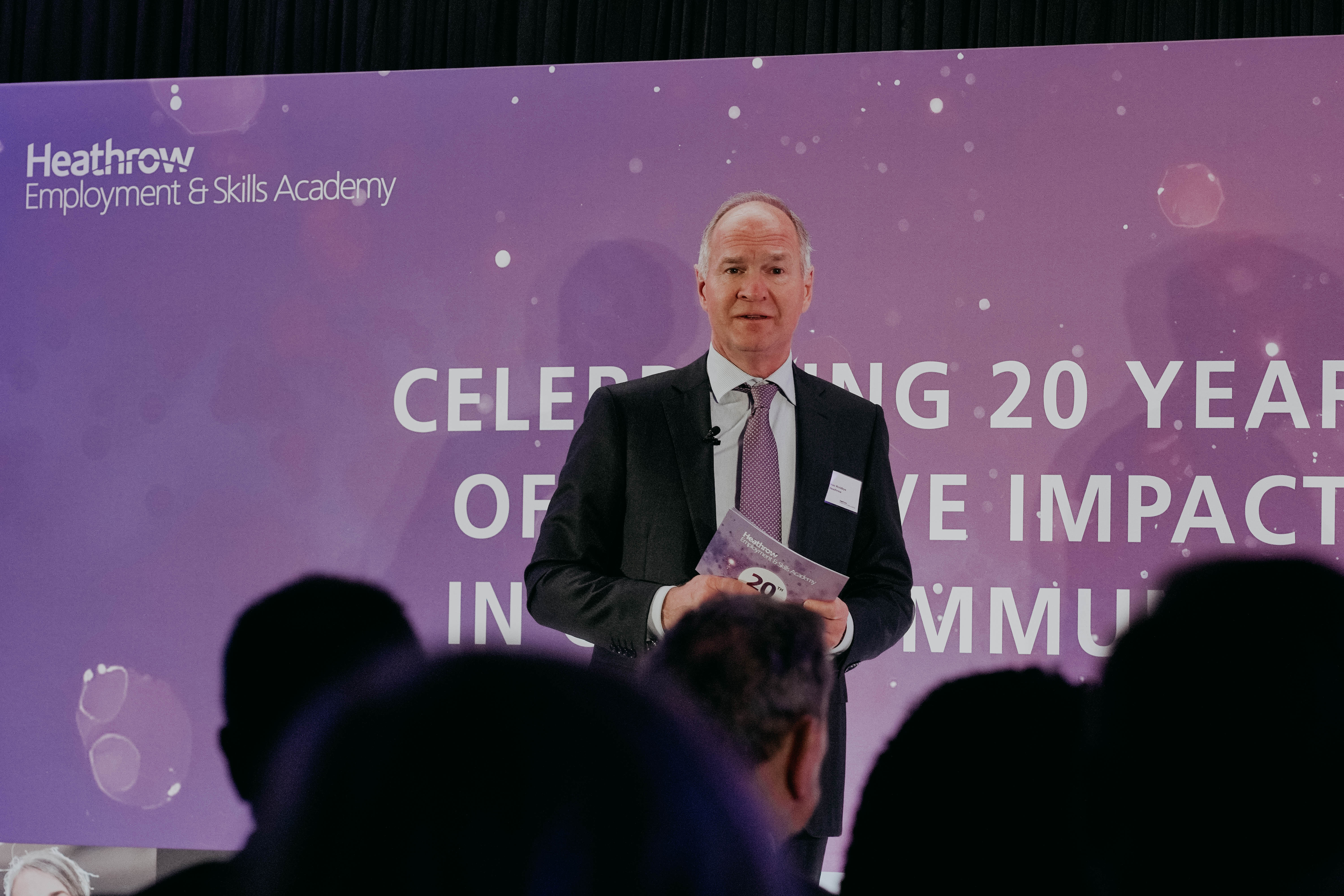 Heathrow CEO Thomas Woldbye speaks on stage in front of a large purple backdrop that reads, "Celebrating 20 years of positive impact in our community." He is viewed from the audience.