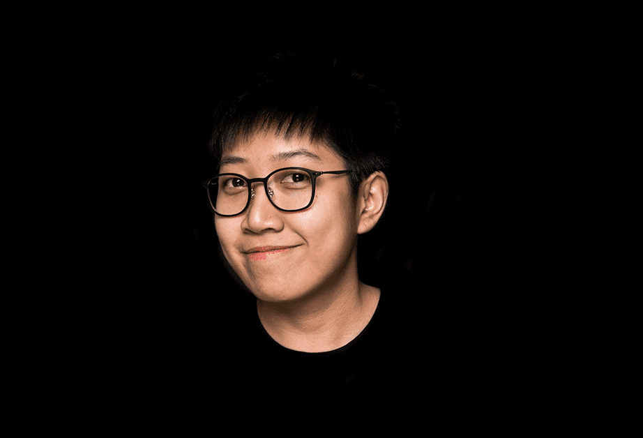 A person with short dark hair and glasses is looking slightly to their right against a solid black background. They are wearing a dark-colored shirt and have a subtle smile.