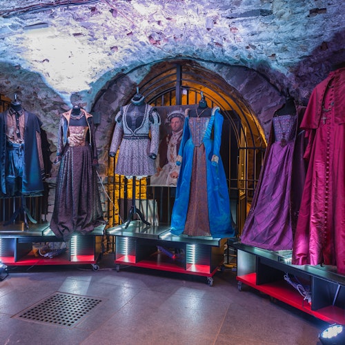 Mannequins display ornate historical costumes in a dimly lit stone room, with an arched doorway and barred gate in the background.