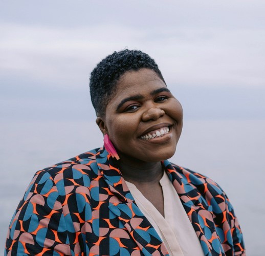 A person is smiling at the camera, wearing a colorful patterned jacket, with a cloudy sky and water in the background.