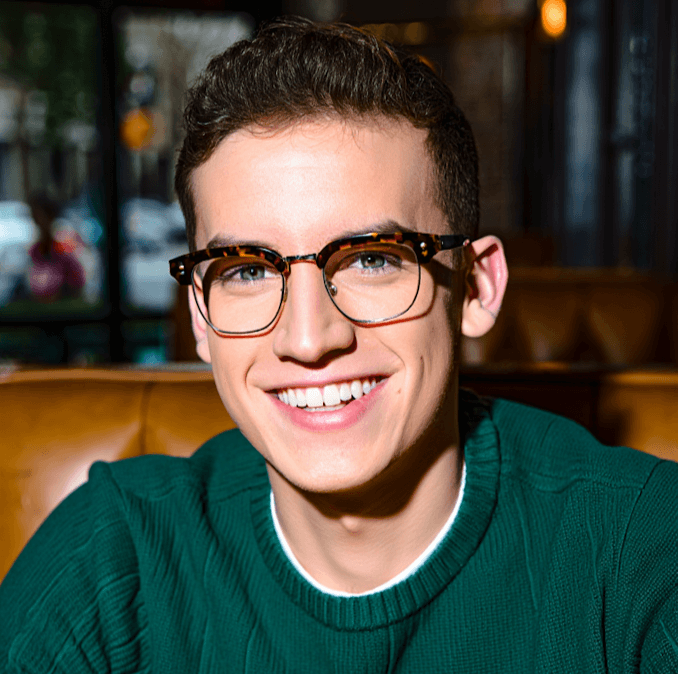 A man wearing glasses smiles softly, his eyes focused on the viewer, conveying a friendly demeanor.