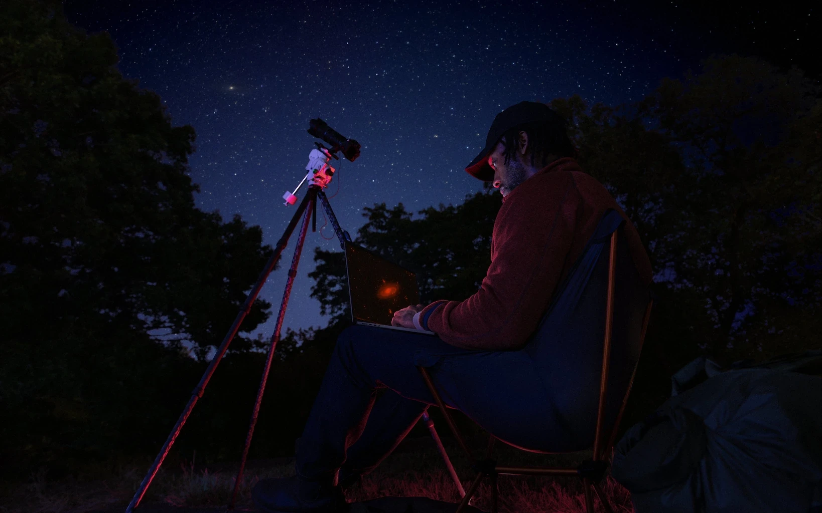 Man sitting outside at night looking at laptop next to star tracker