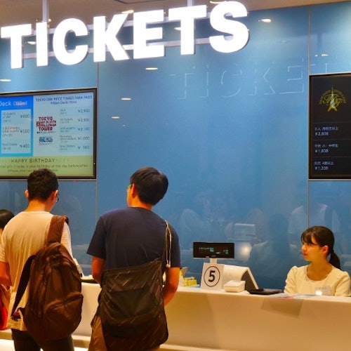 三個人站在票務櫃檯，櫃員在一個大型的燈光閃爍的「票」標誌下。數碼屏幕顯示票價。