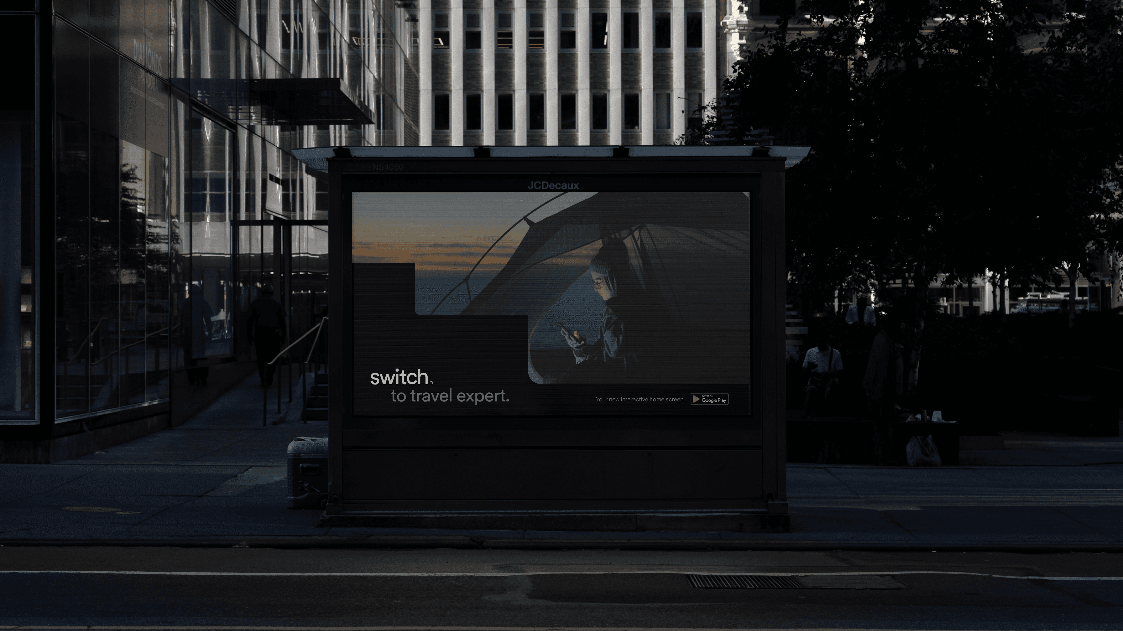 A dark-colored bus stop displays a large advertisement featuring a silhouetted person holding an umbrella against a sunset backdrop. The ad includes the text "switch. to travel expert." and "Your next immersive experience is PG." The bus stop is situated on a sidewalk beside a street, with tall buildings visible in the background and trees to the right.