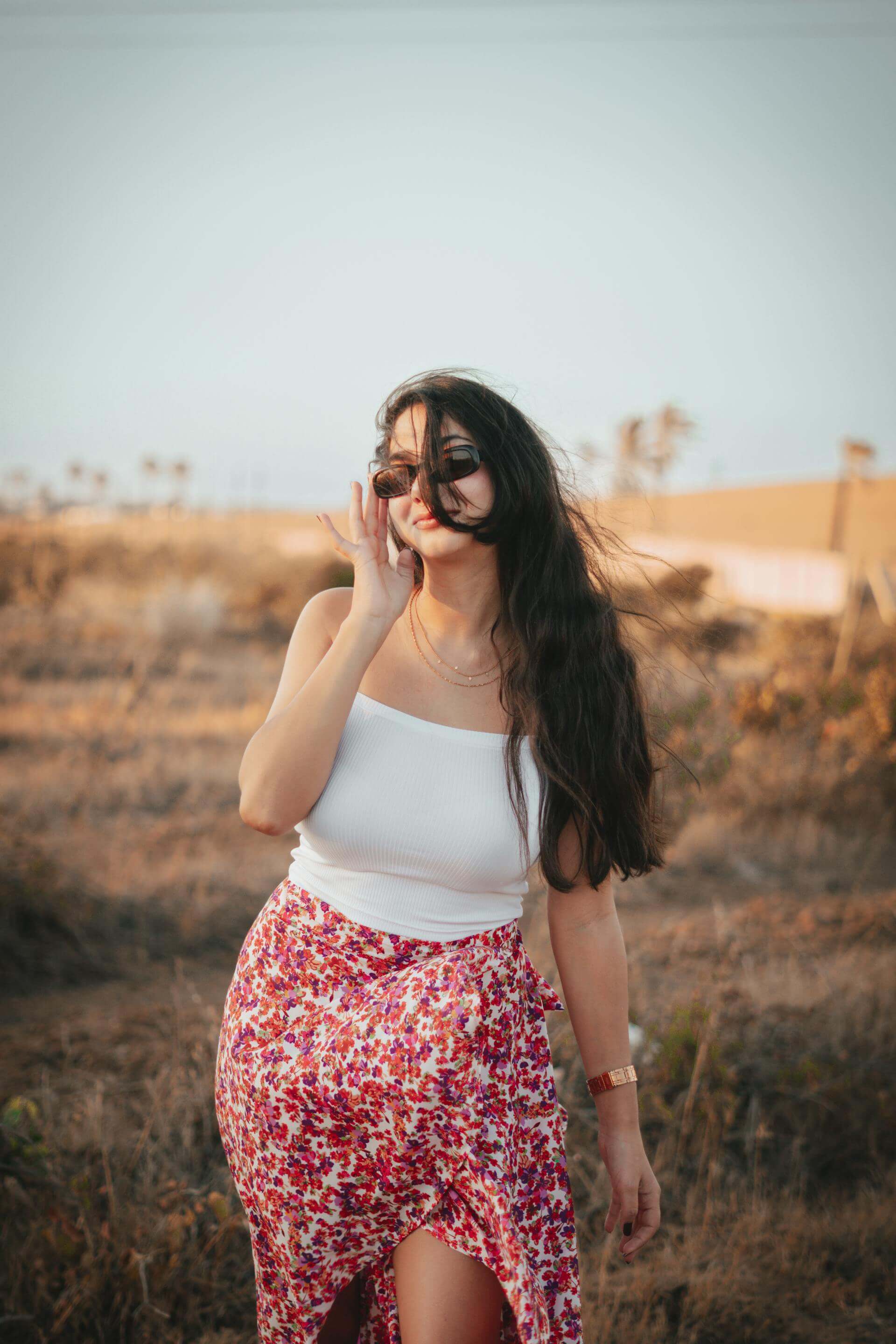 A woman in a floral skirt and white top strolls through a vast desert landscape under a clear blue sky