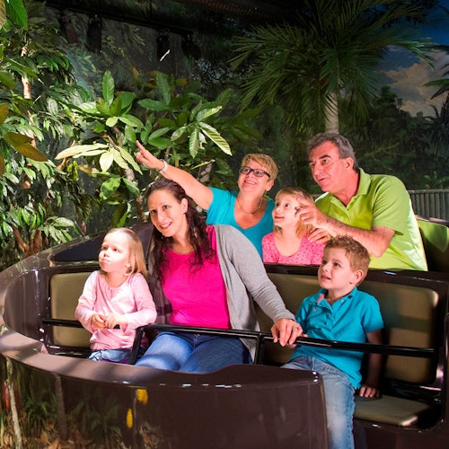 A family of six, including three children, rides an amusement park boat surrounded by lush, jungle-like vegetation.