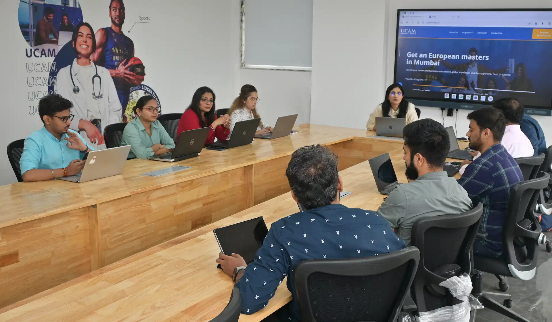A diverse group of individuals sits around a large wooden conference table, engaged in a meeting; laptops are open, with a presentation visible on a screen showing information about higher education opportunities at UCAM Mumbai University.