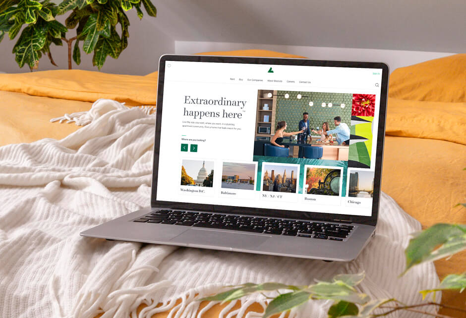 A laptop on a bed displays a website with travel destinations and photos. A blanket and green plants are visible in the background.