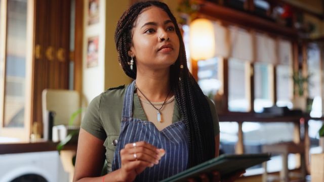Mulher preta de tranças longas e avental listrado segurando um tablet em ambiente de cafeteria.