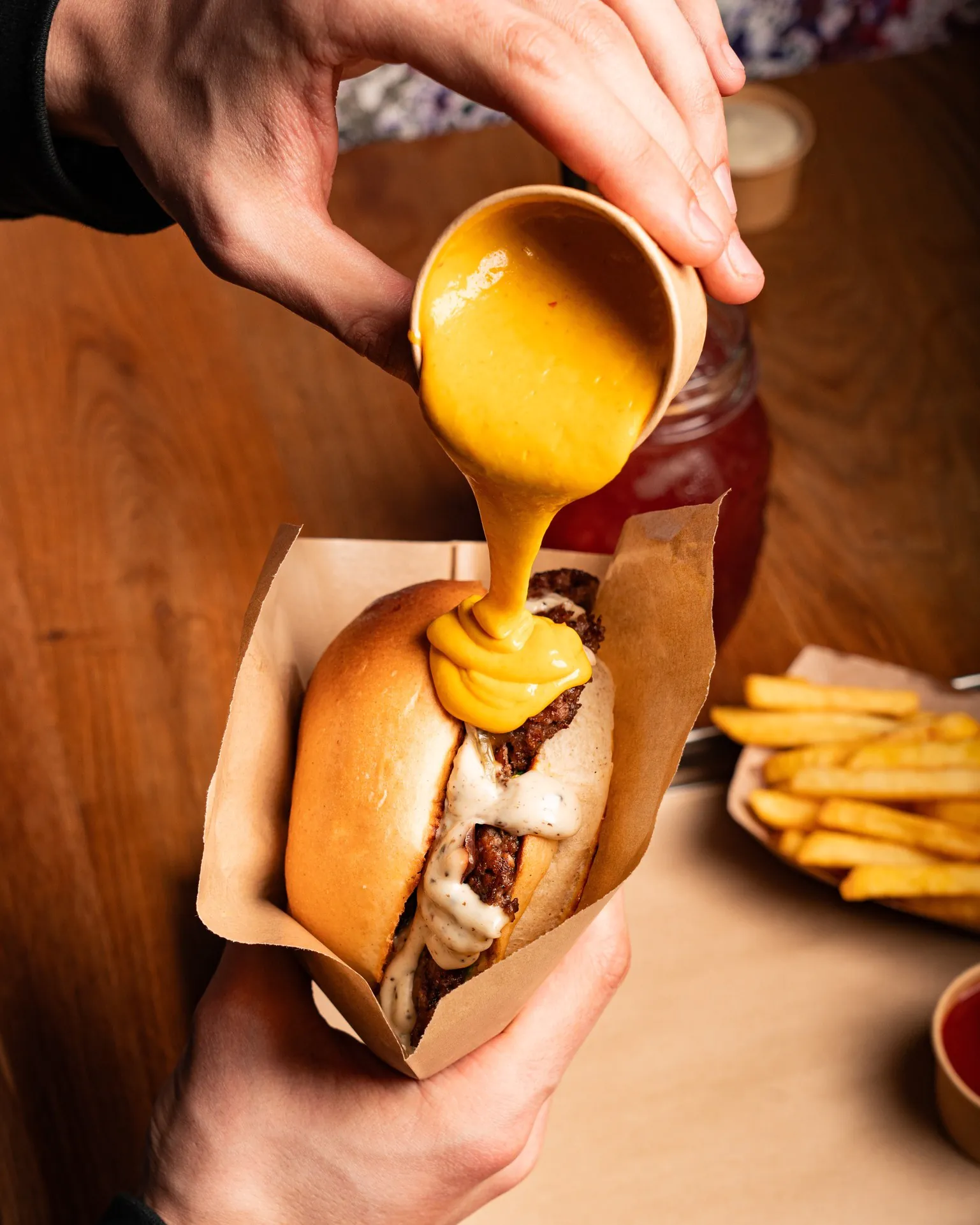 A person pours mustard over a burger, with fries visible in the background.