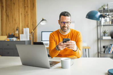 A person sat at in a home office with their laptop on the desk but also looking at their phone with keen interest