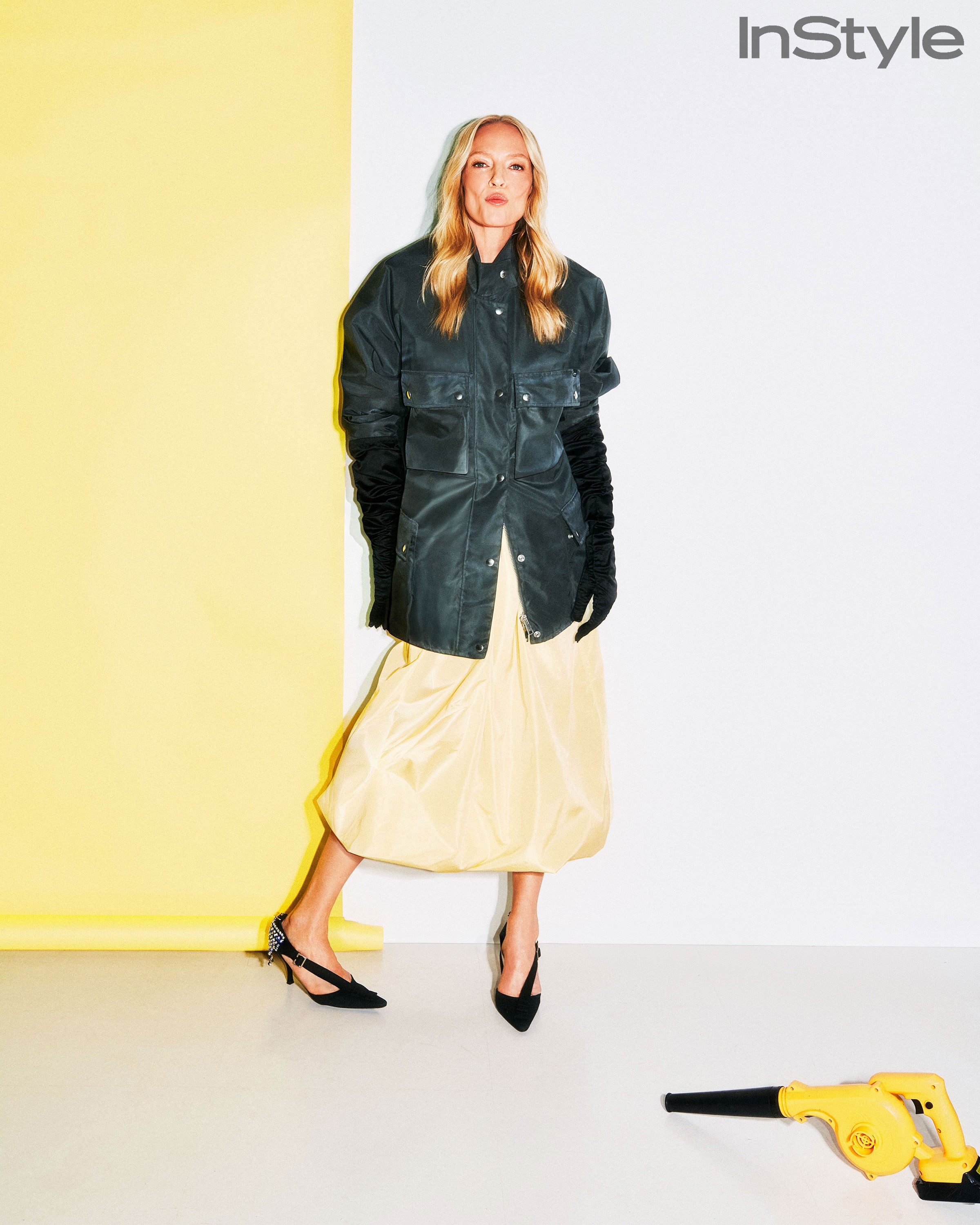 Uma Thurman standing in a black jacket and pale yellow skirt beside a yellow set wall