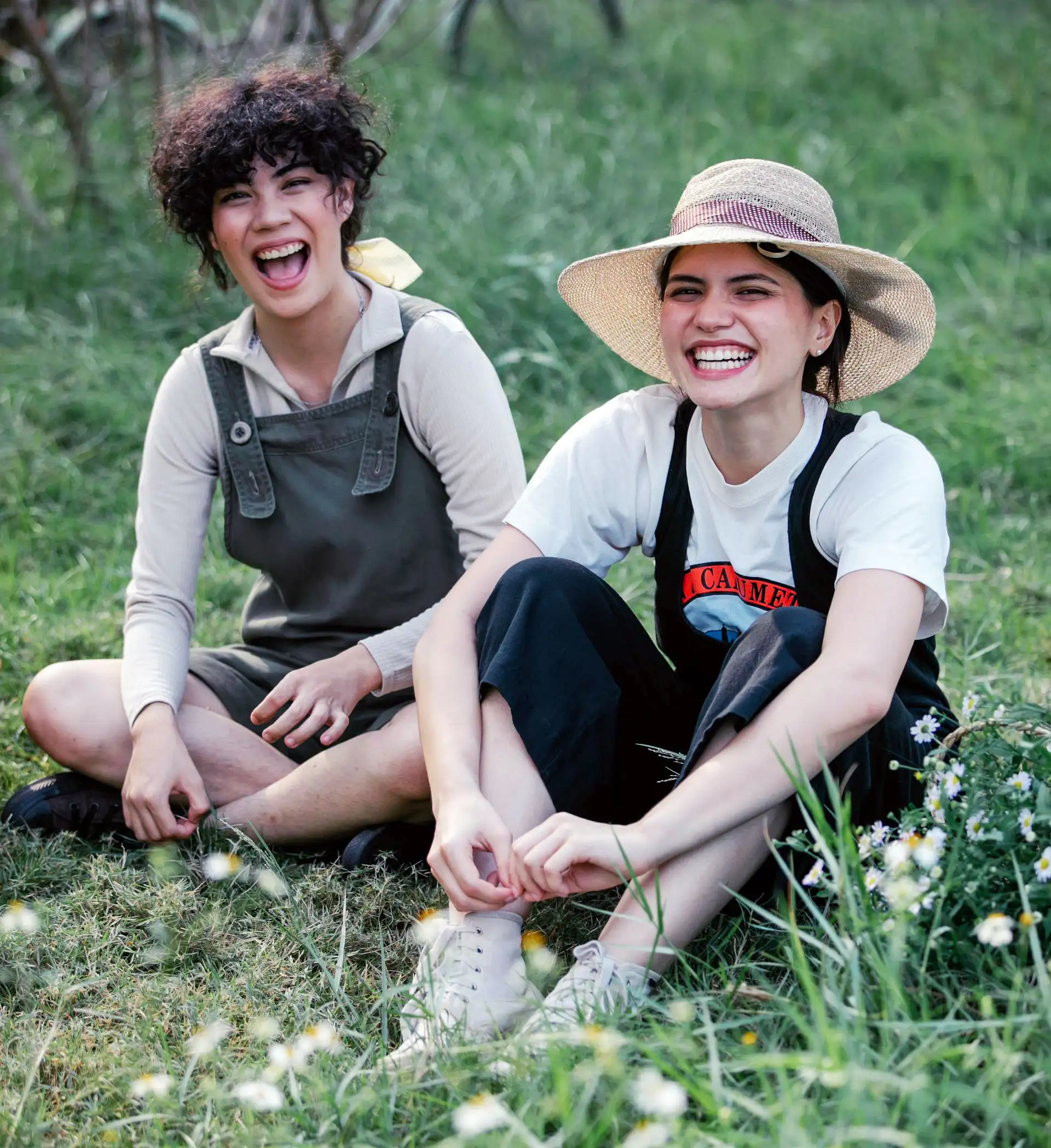 Two laughing young women in overalls and sun hats sit in a grassy field.