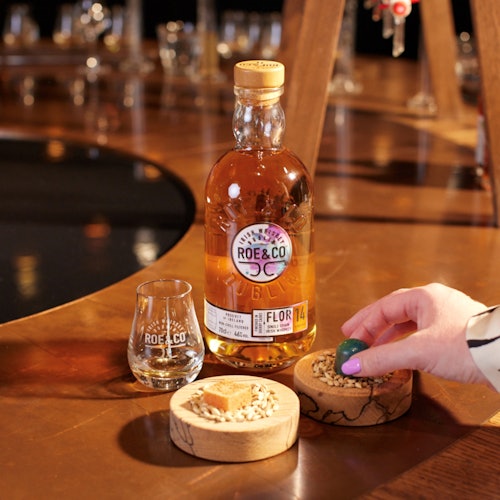A bottle of Roe & Co whiskey beside a branded glass and two wooden dishes, one with a sugar cube and another with seeds, placed on a wooden table.