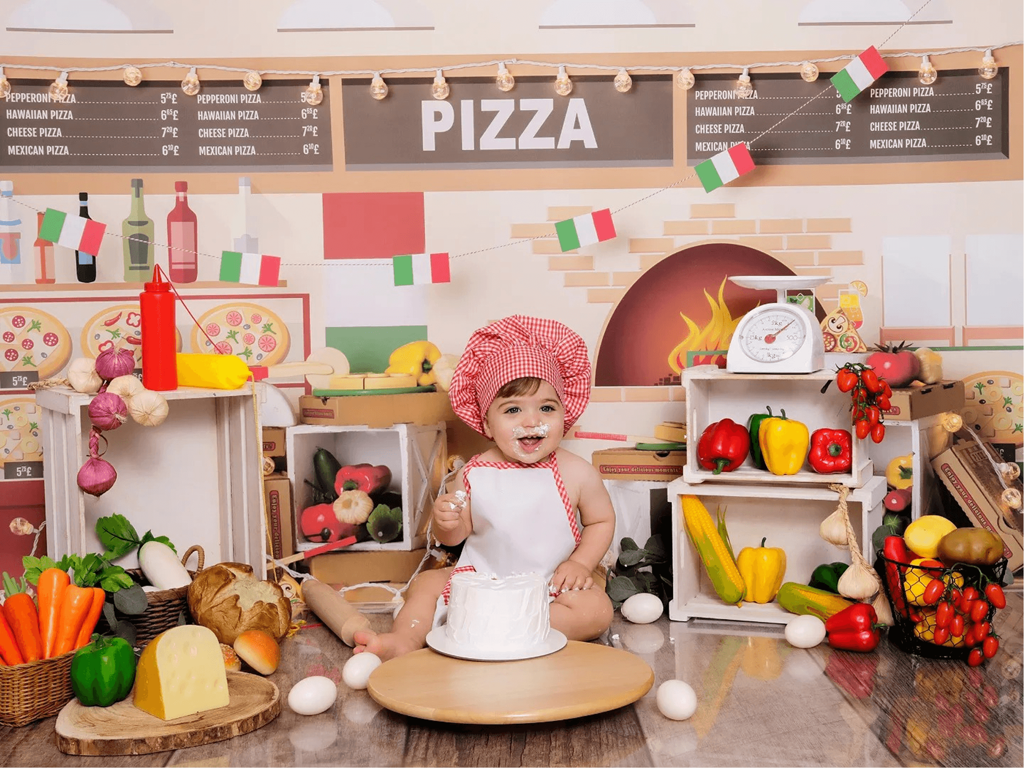 A baby is in a chef outfit, sitting in a pizza-themed, monthly baby photo idea. There are vegetables, eggs, and an Italian pizza shop background. 