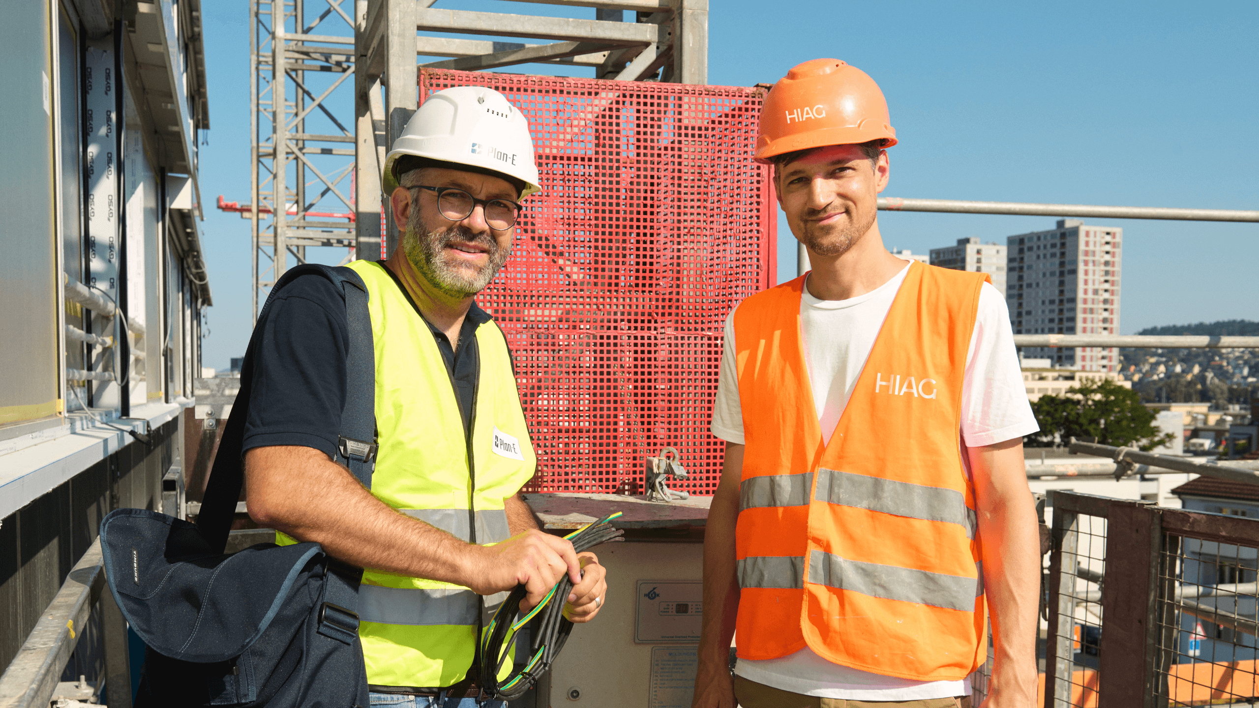 Zwei Fachpersonen auf der Baustelle des ALTO-Towers mit Schutzhelmen und Warnwesten, im Austausch vor installierter Gebäudetechnik in luftiger Höhe.