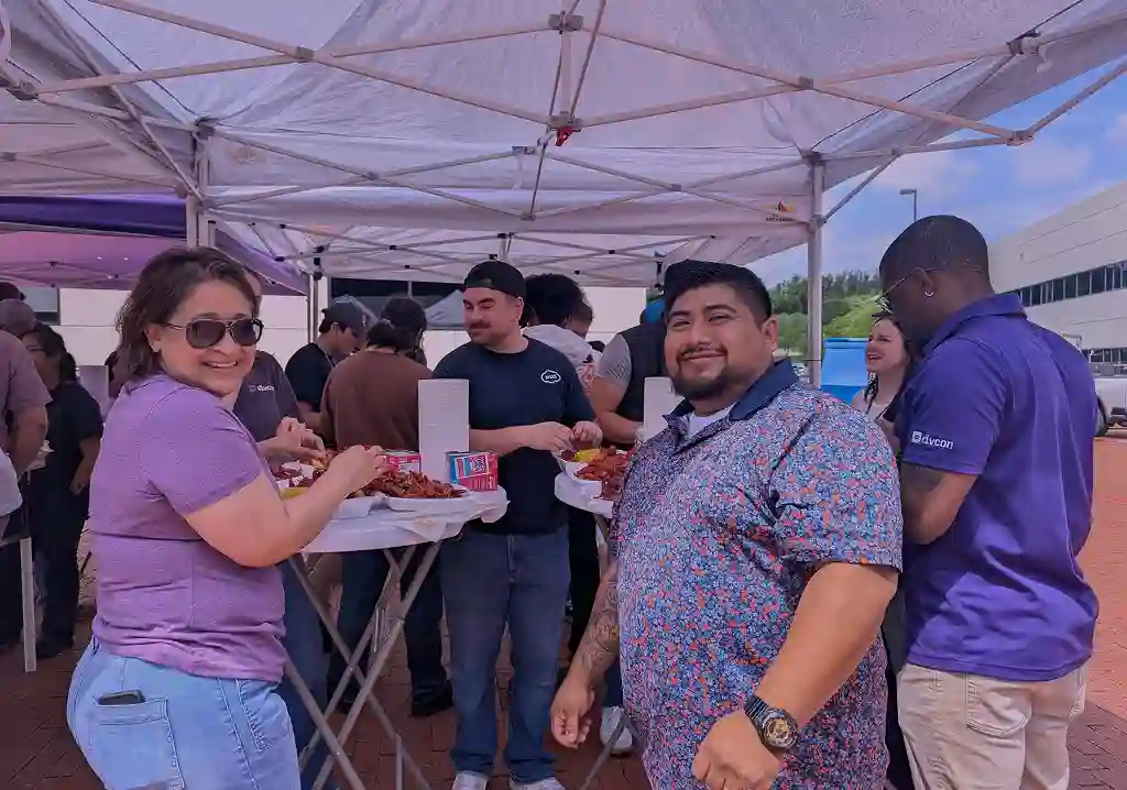 People gathered outdoors under white pop-up tents, standing around high-top tables enjoying a crawfish boil. In the foreground, a woman wearing sunglasses and a purple shirt smiles while getting food from a plate, beside a man in a patterned blue shirt smiling at the camera.