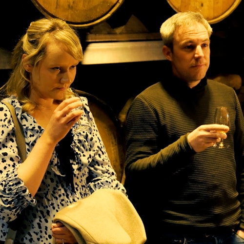 Two people are sampling drinks in front of wooden barrels. The woman holds a glass and a coat, while the man holds his glass and looks ahead.