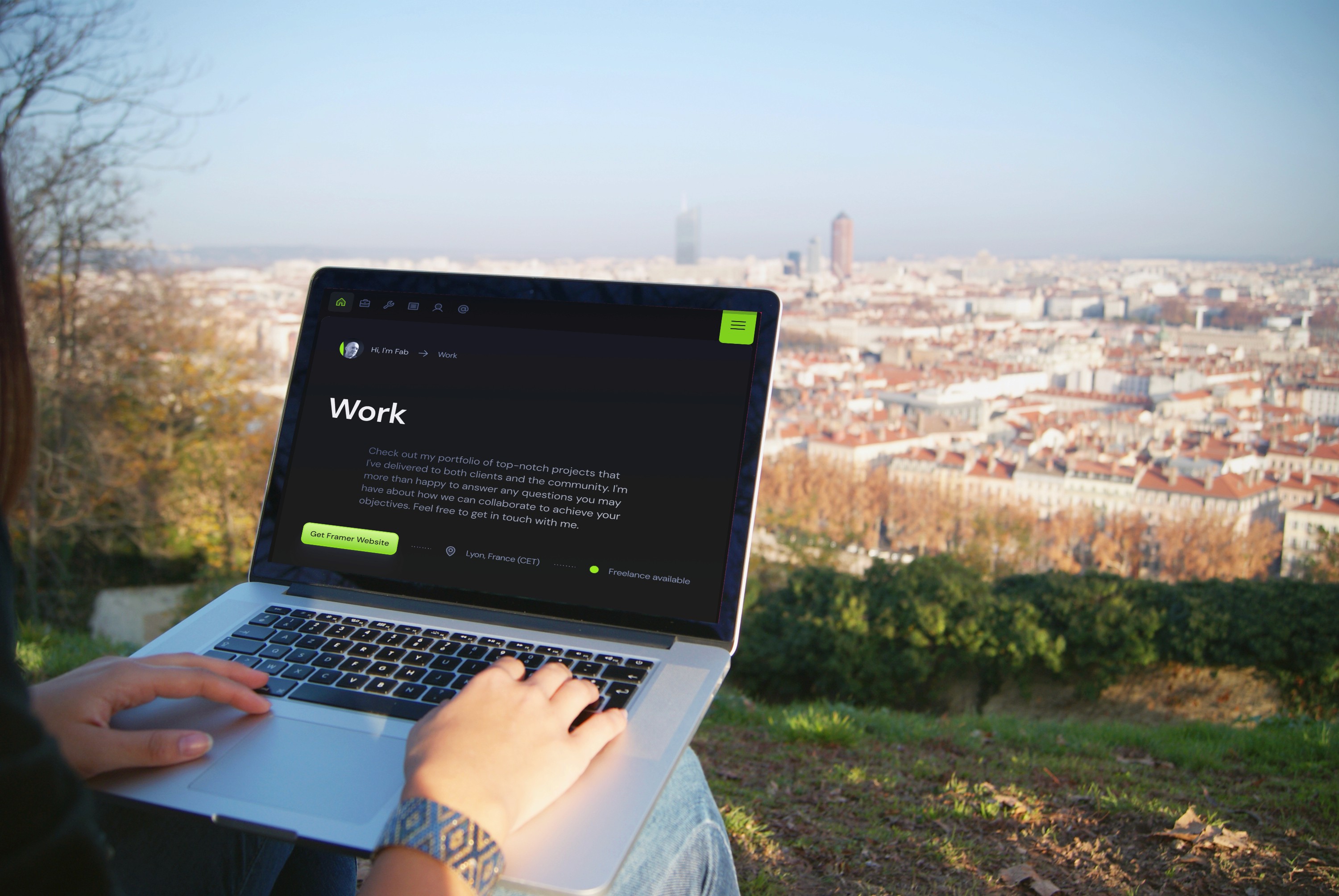A person using a laptop displaying a webpage titled "Work" with a blurred cityscape visible in the background from a grassy hilltop.
