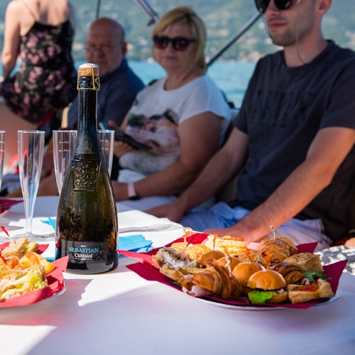 People sitting at a table on a boat with a bottle of wine, glasses, sandwiches, and croissants, with a scenic water view in the background.
