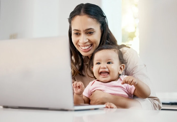 A imagem mostra uma mulher sorridente segurando um bebê em seu colo enquanto ambos olham para a tela de um laptop. O bebê, que veste uma camiseta rosa, também está sorrindo, demonstrando alegria e entusiasmo. A mulher parece estar trabalhando ou fazendo uma videochamada, mas claramente está envolvida em um momento feliz com seu filho. A atmosfera é leve e acolhedora, e a cena retrata a conciliação entre o trabalho remoto e os cuidados maternos, destacando a conexão e o carinho entre mãe e filho.