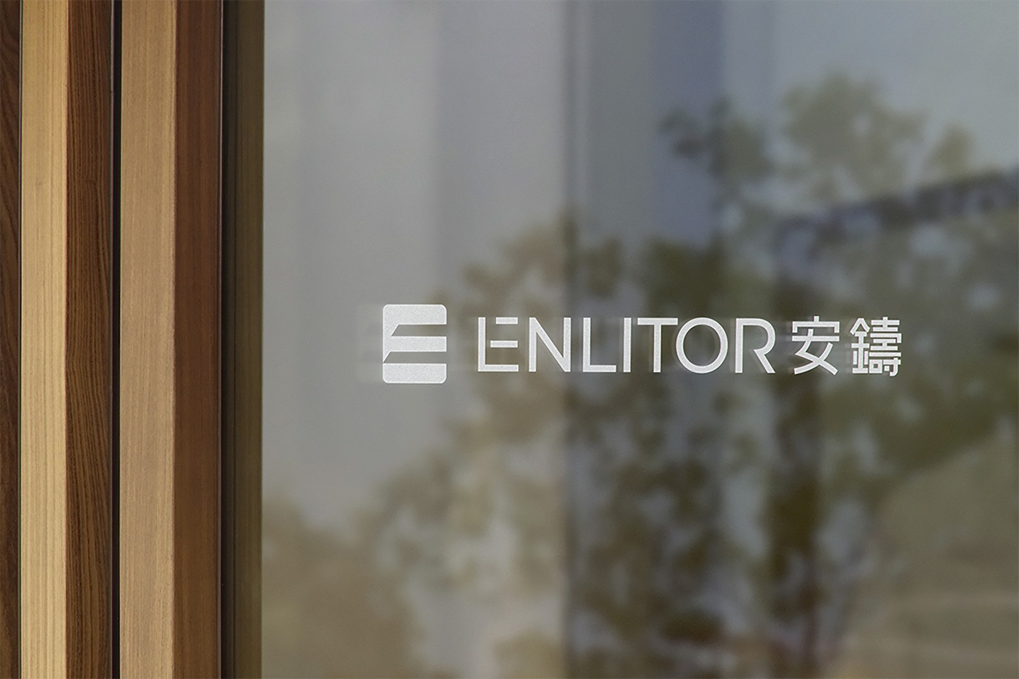 這張圖片展示了Enlitor的品牌識別設計。標識貼在透明的玻璃上,背景模糊,突出標識的簡約設計。