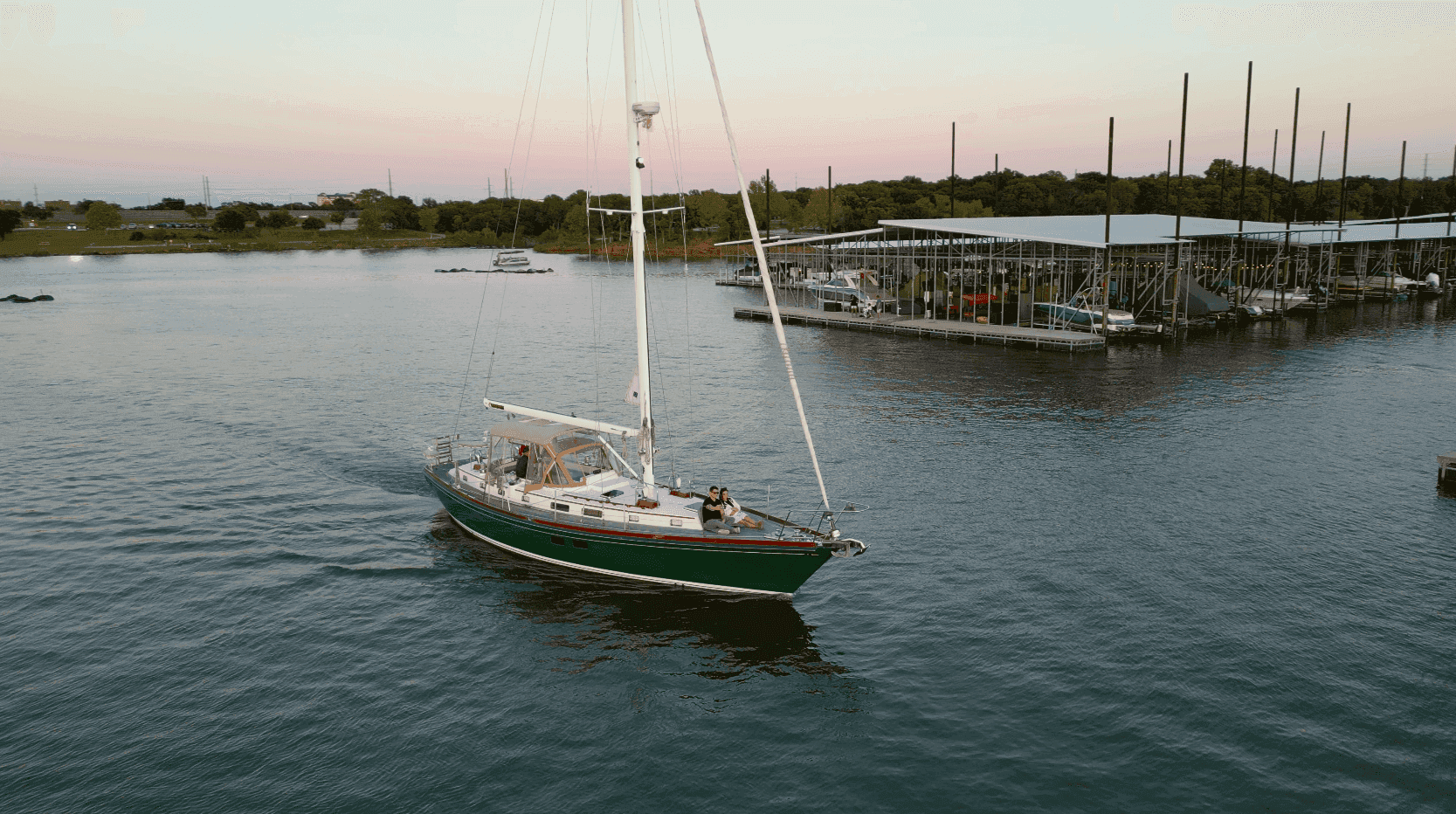 A green sailboat glides across a calm marina, with a backdrop of a dock and trees under a pastel sunset sky.