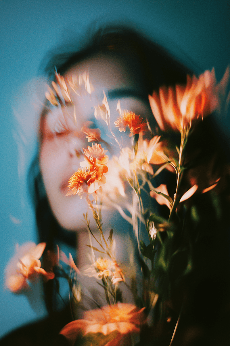 Artistic portrait of a woman partially obscured by orange flowers, captured with soft motion blur and teal-toned lighting.