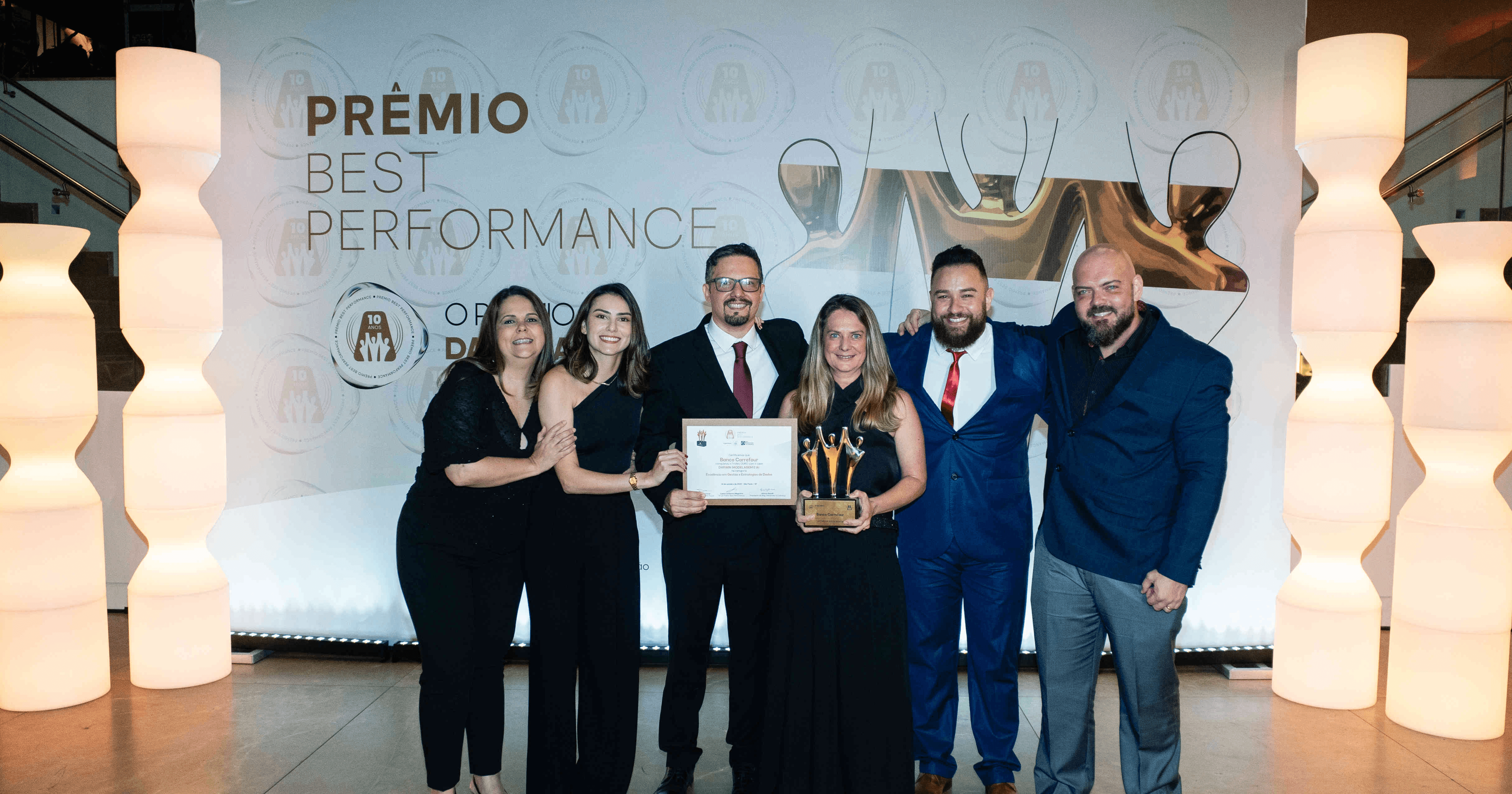 Executivos do Banco Carrefour posando com troféu ouro e certificado no palco do Prêmio Best Performance 2025