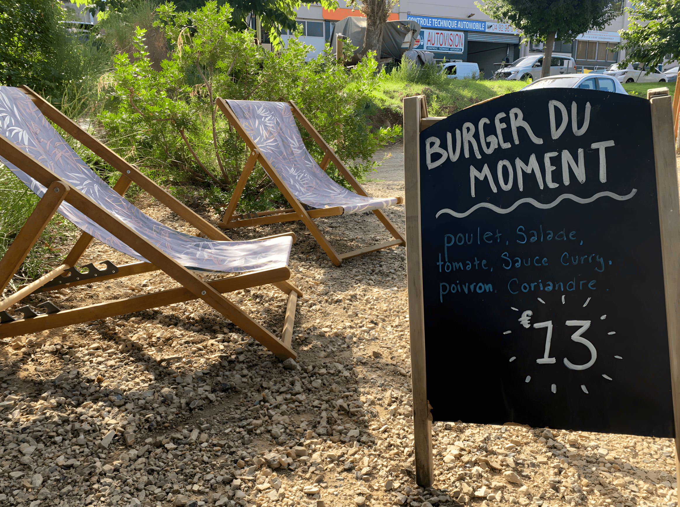 Deux transats sur un fond de buisson avec un chevalet annonçant le burger du moment.