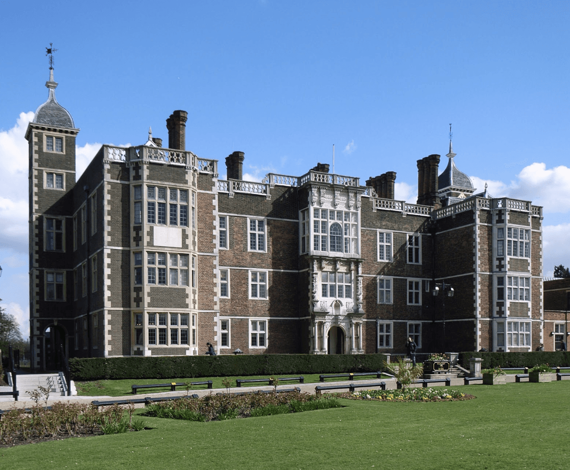 A smiling man with curly hair is sitting at a desk, looking at the camera, with a laptop and a lamp in the background.