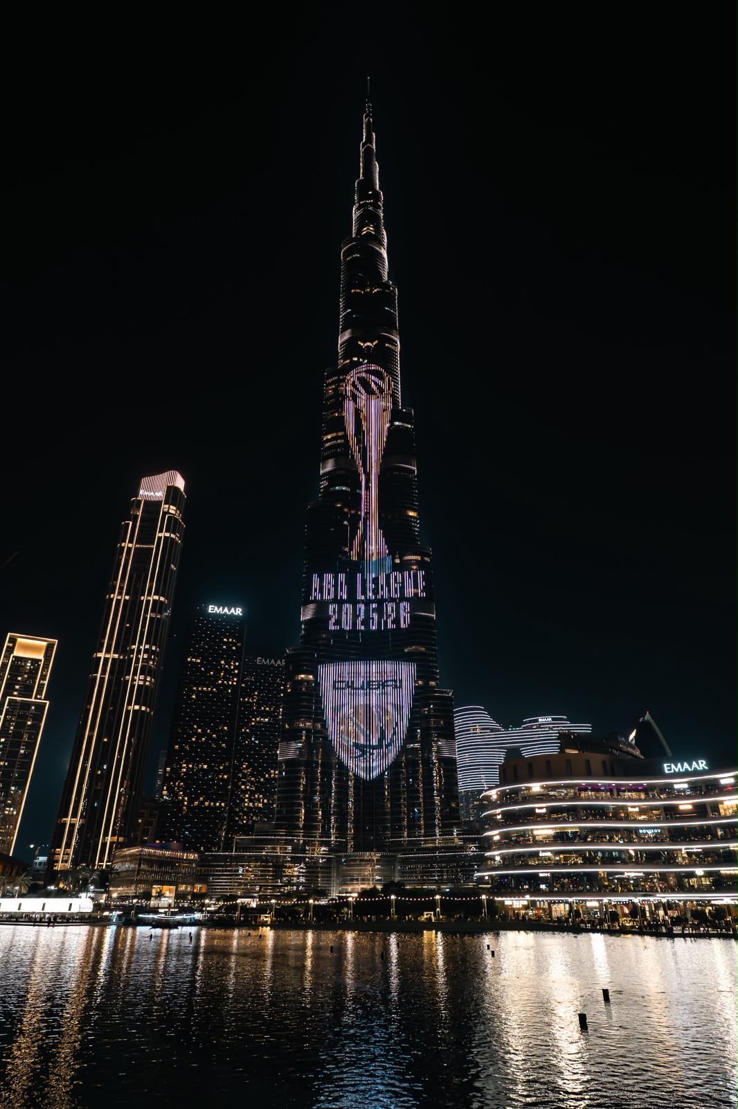 Burj Khalifa illuminated at night with a large projected portrait, overlooking the Dubai Fountain waterfront.