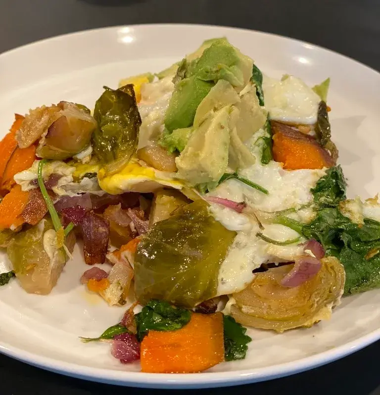 A vibrant salad featuring roasted vegetables, including Brussels sprouts and sweet potatoes, accented with creamy avocado slices, red onion, and fresh greens on a white plate.