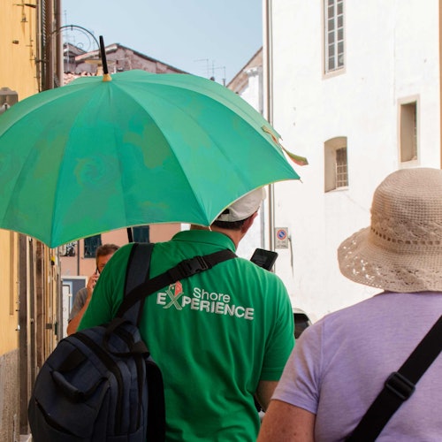 Due persone camminano lungo una strada soleggiata, una tiene un grande ombrello verde e indossa una maglietta verde con la scritta "Esperienza Costiera."