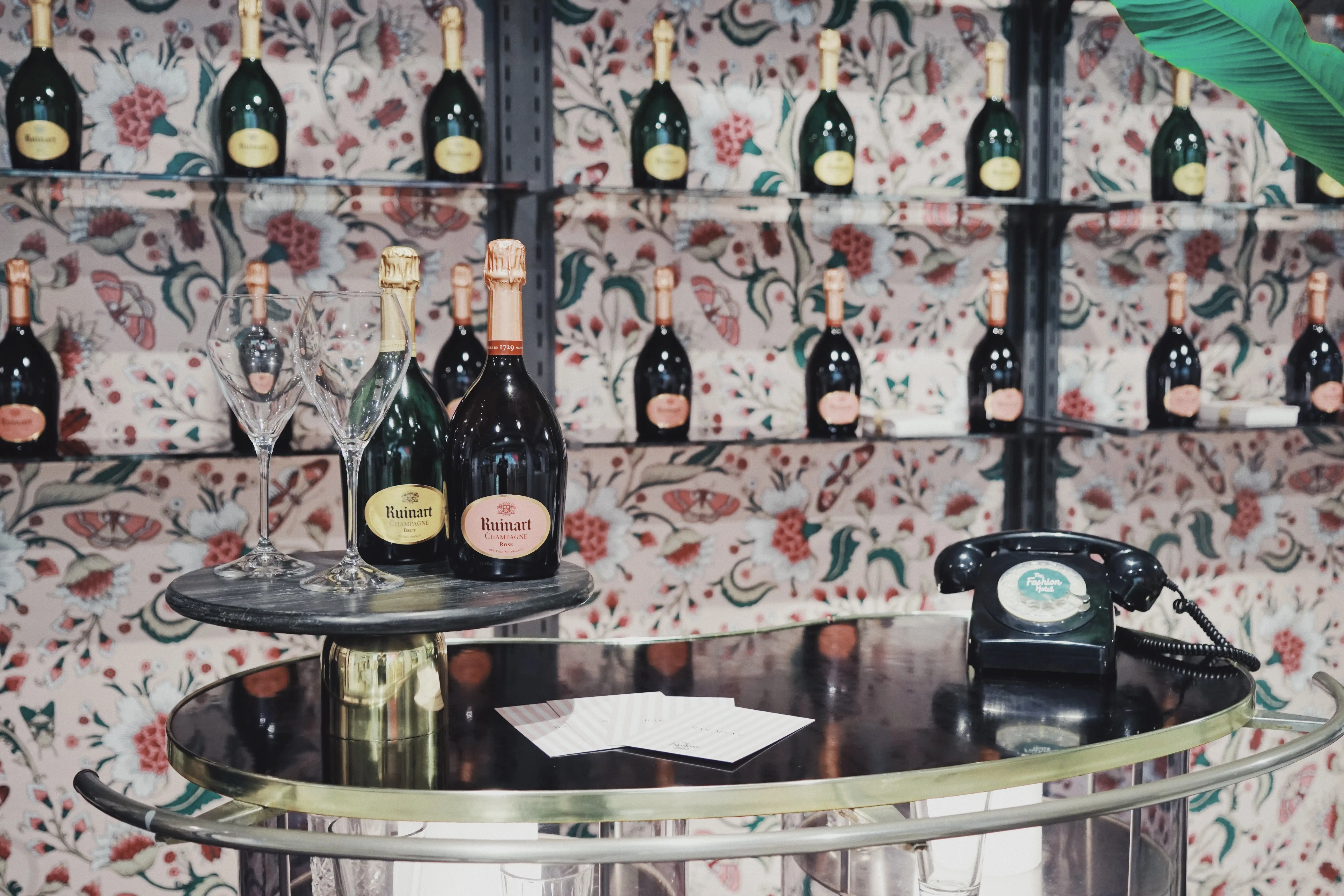 Champagne bar detail at UZWEI FASHION HOTEL with Ruinart bottles on a mirrored trolley, a vintage black telephone and floral wallpaper shelves lined with more bottles.