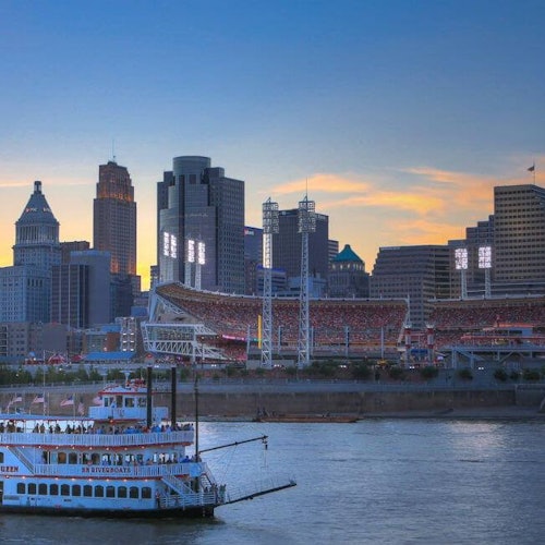 日落時分的城市天際線景觀，前景有一艘河船，背景有一座燈火通明的體育場。