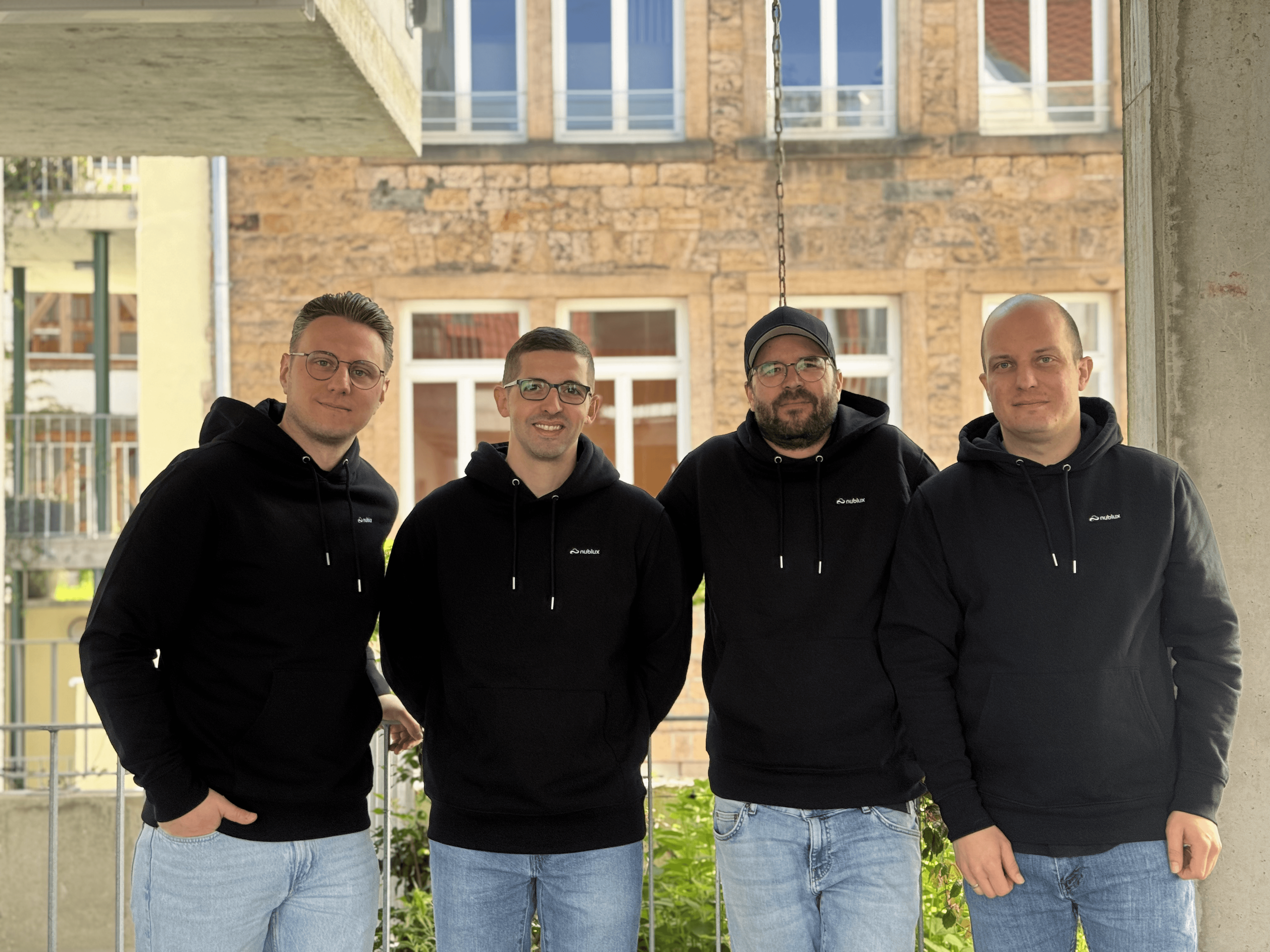 A young person with curly hair smiles at the camera while sitting in front of a laptop against a dark background.