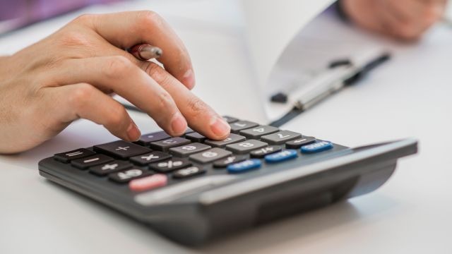Mão segurando caneta entre os dedos, digitando em calculadora sobre a mesa