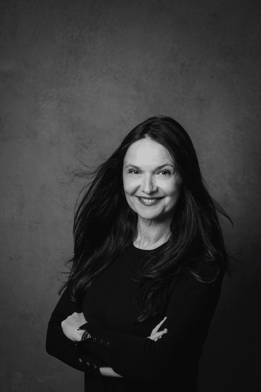 Porträt einer lächelnden Frau mit braunen Haaren, verschränkten Armen und einem schwarzen Oberteil.