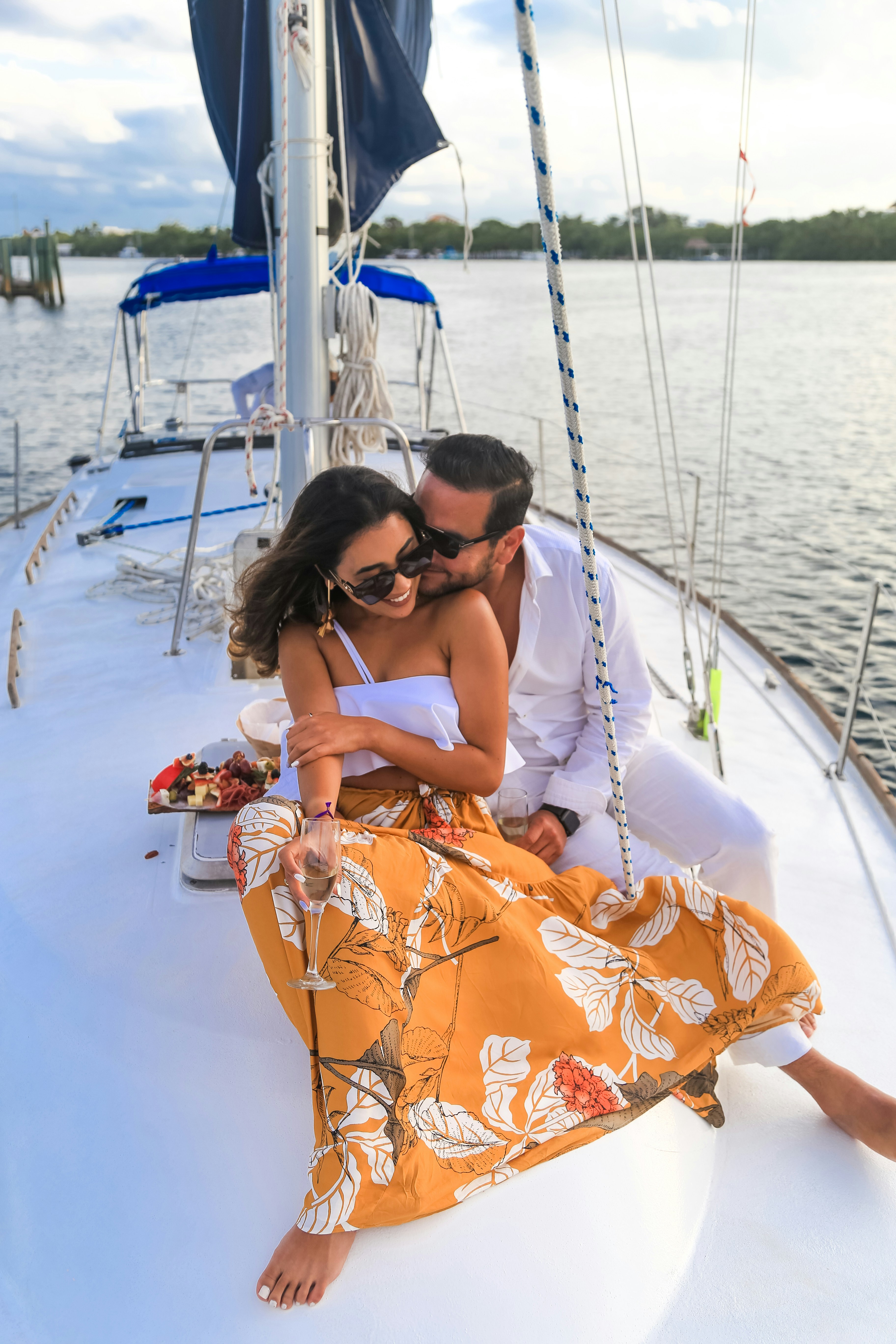 A couple enjoying a romantic moment on the deck of a sailboat, with the woman in a floral orange skirt holding a glass of champagne, set against a tranquil water landscape.