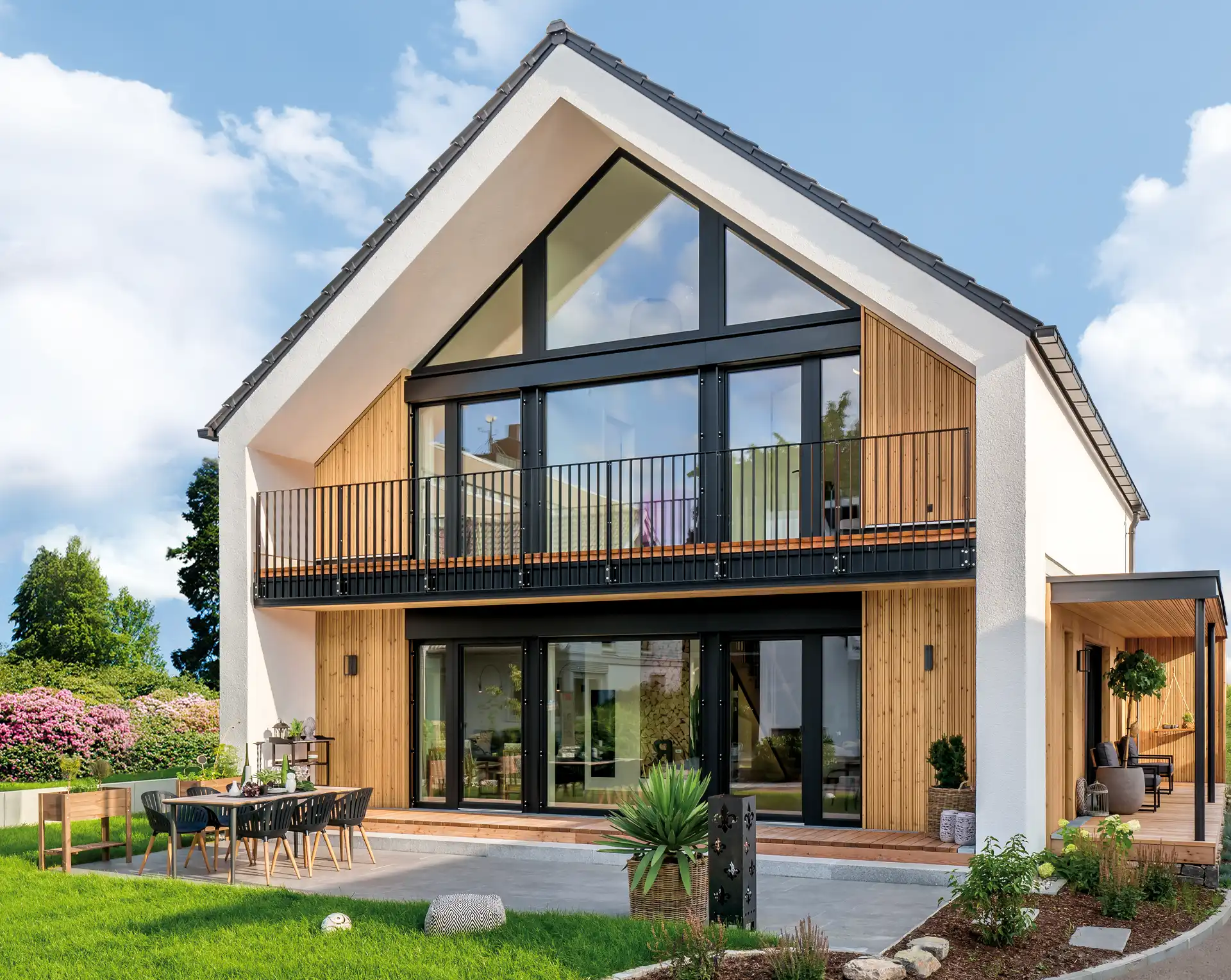 Modernes Einfamilienhaus mit großer Fensterfront, Holzakzenten und einem Balkon im Sommer.