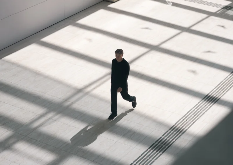 Felix Maier in dunkler Kleidung geht durch lichtdurchflutete, moderne Halle mit geometrischen Schattenmustern – urbanes Setting und minimalistisches Architektur-Design.