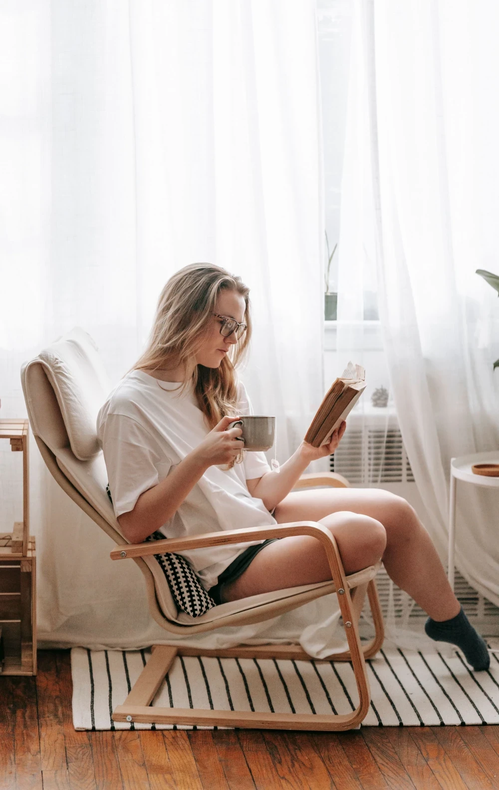 Woman relaxing in a chair by a bright window, reading a book and holding a mug.