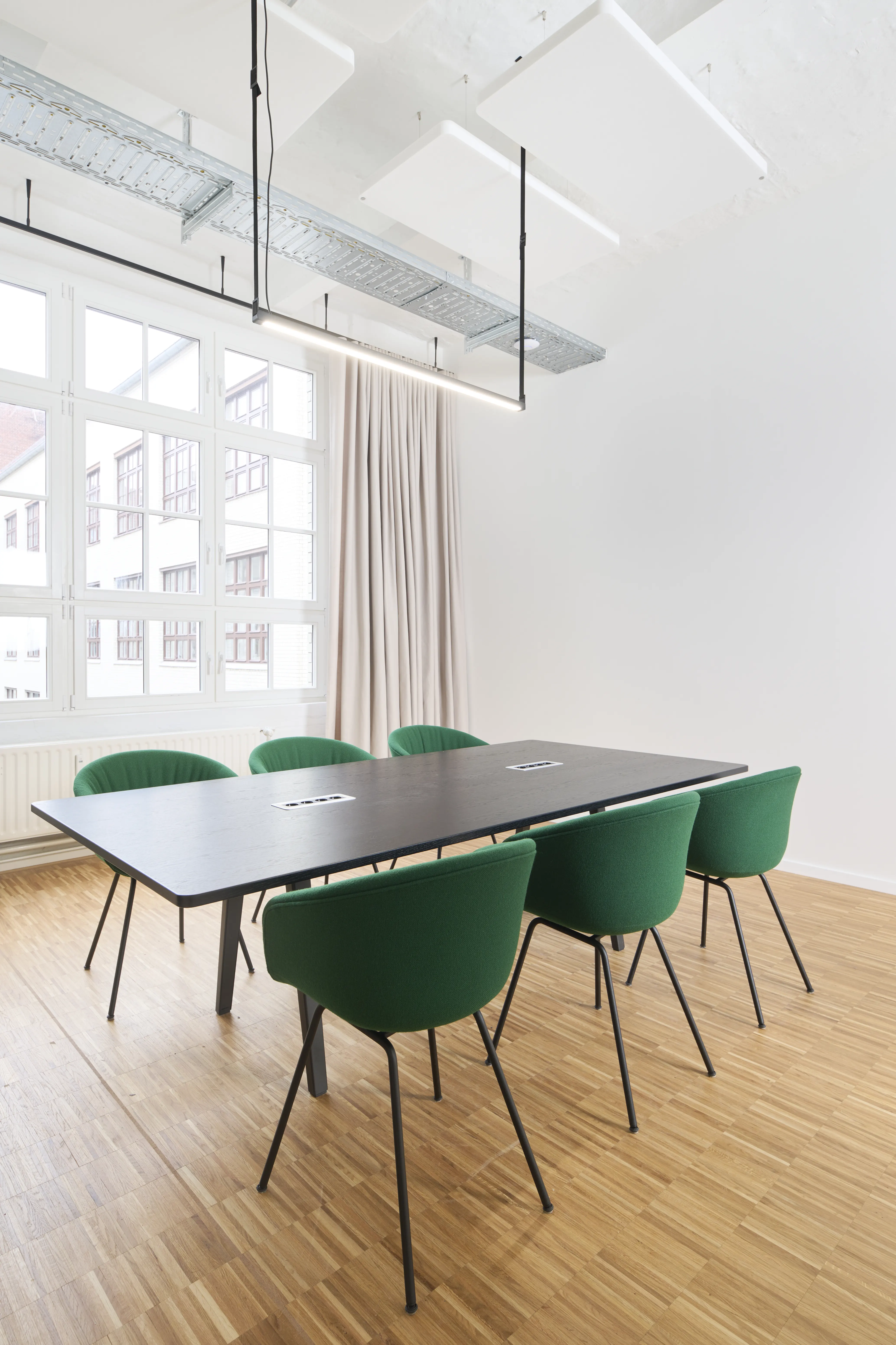 Meeting room at PLAN A with a black table, six emerald-green upholstered shell chairs, a black rail-mounted linear light hanging from the exposed ceiling and beige curtains by the window.