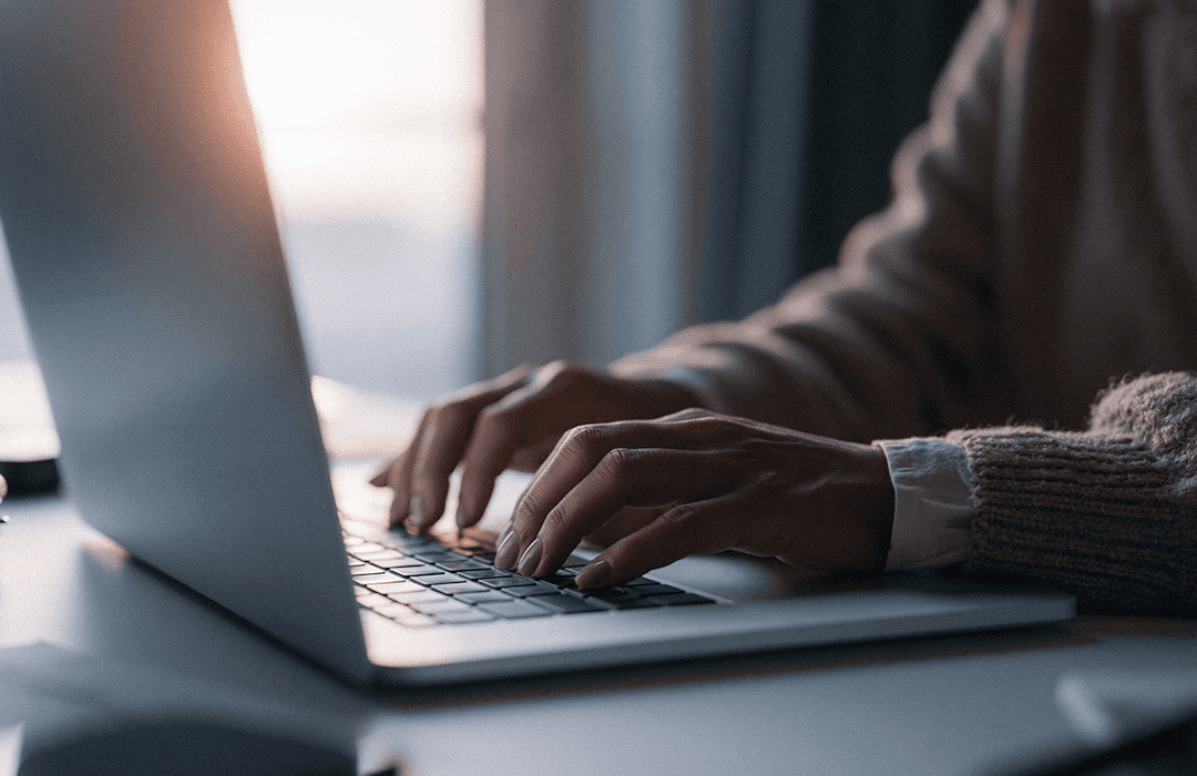 Close-up focado nas mãos de uma pessoa digitando no teclado de um notebook prata sobre uma mesa. O ambiente tem iluminação suave e acolhedora, vinda do fundo, transmitindo uma atmosfera de trabalho focado ou escrita de artigos.