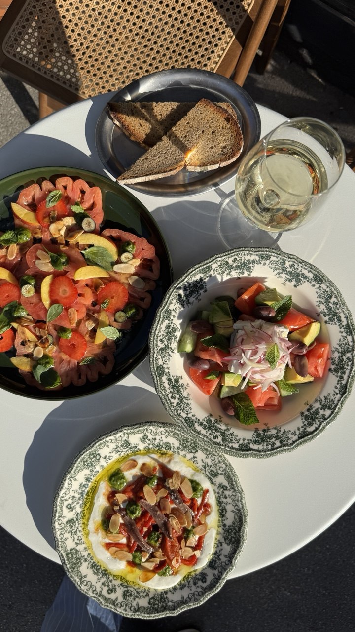 A sunlit outdoor table features a vibrant spread with a bowl of sliced tomatoes topped with herbs, a plate of mixed salad with tomatoes, cucumbers, and onions, a dish of creamy dip garnished with nuts and herbs, complemented by rustic bread slices and a glass of white wine.