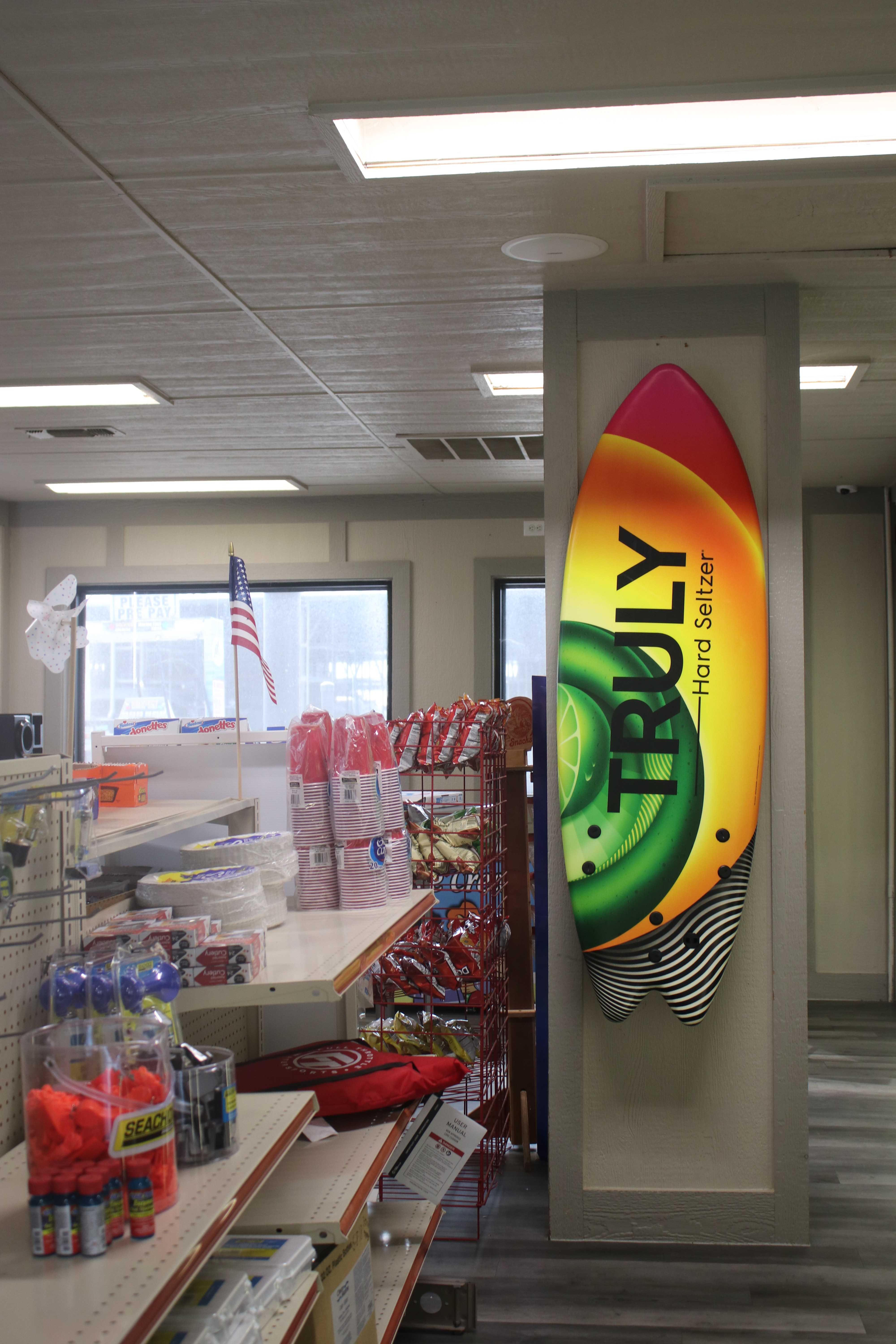 Interior of a convenience store featuring a vibrant "Truly Hard Seltzer" surfboard-style sign on the wall, with shelves stocked with various snacks and goods, and an American flag in the background.