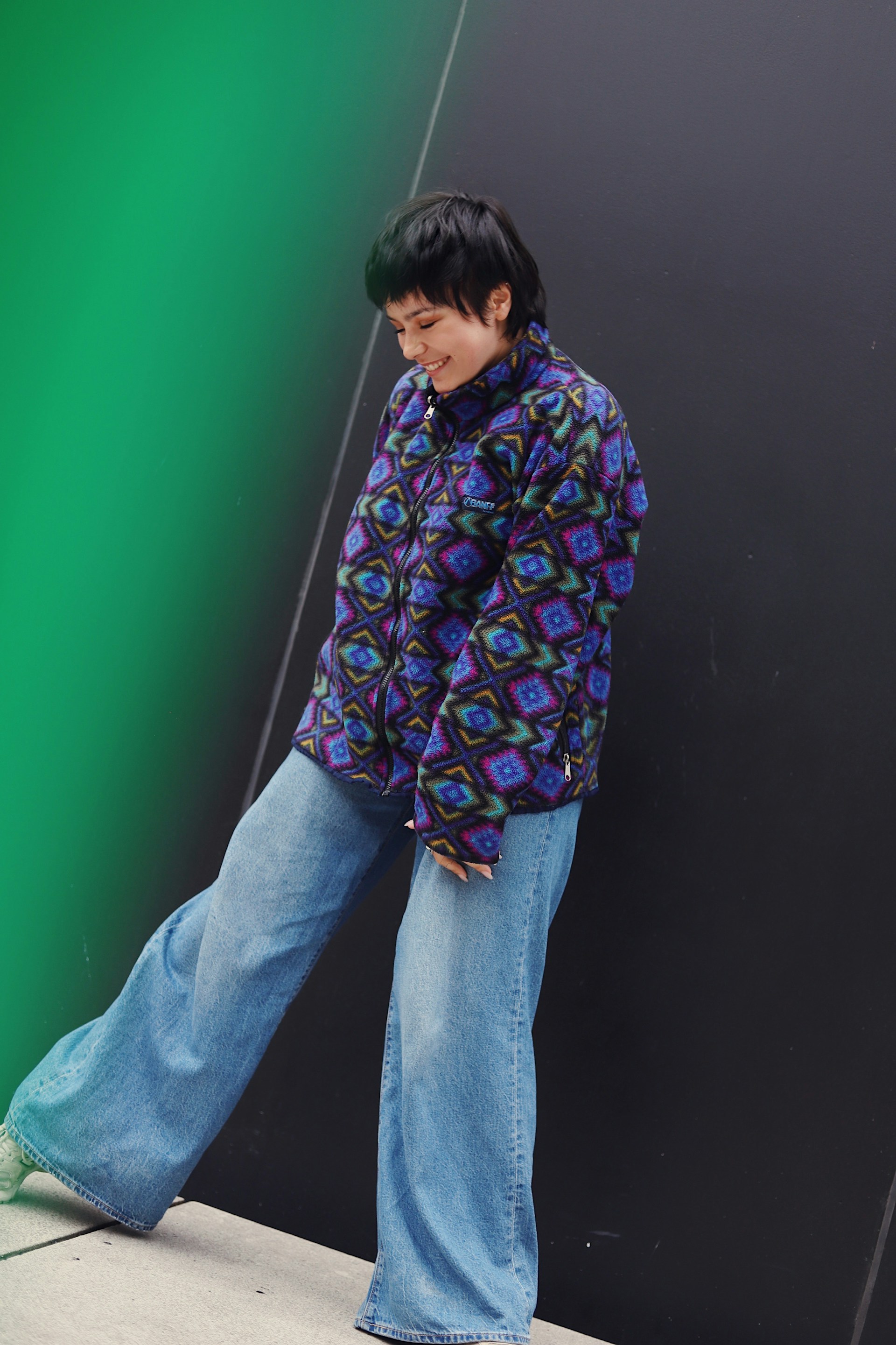 A full-body shot of the same person smiling and looking down while standing against a black wall with a bright green vertical light. They are wearing the patterned fleece jacket and wide-leg blue jeans.