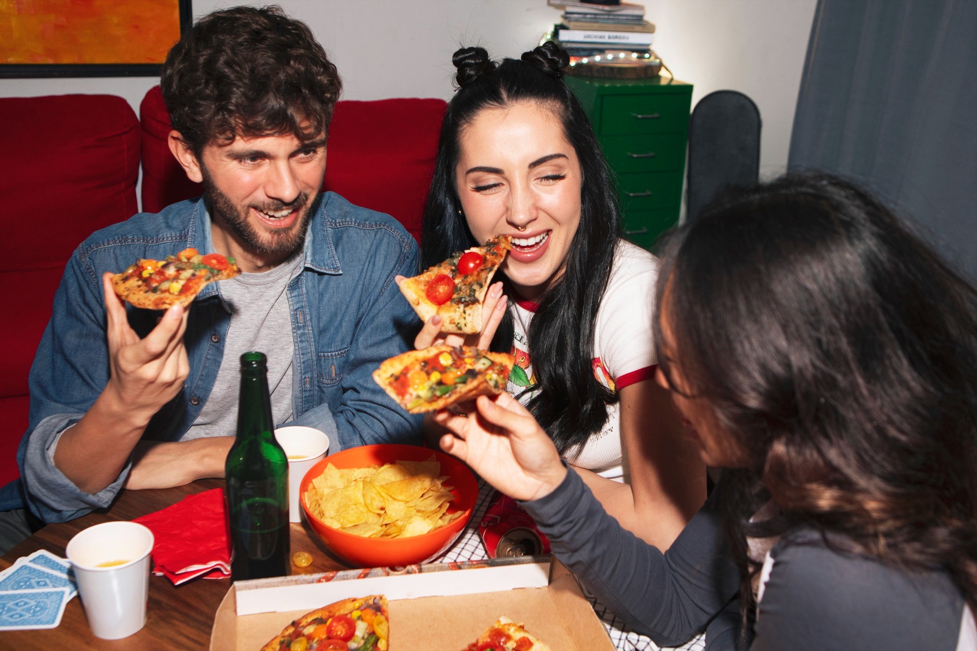 Three smiling friends sitting around a table, laughing and eating slices of pizza together.