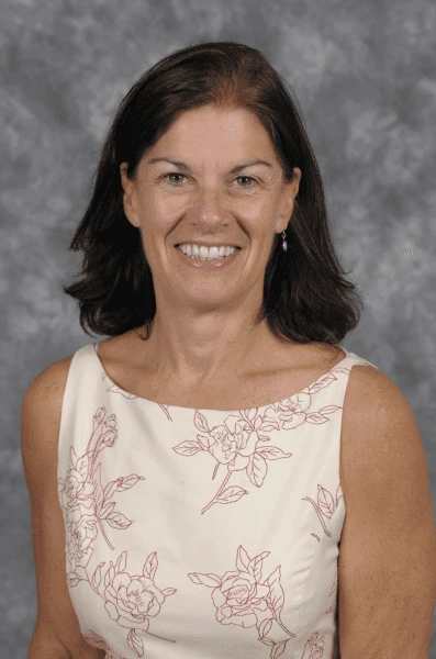 A smiling woman with shoulder-length brown hair wears a sleeveless, white dress adorned with pink floral patterns, standing against a gray marbled background.